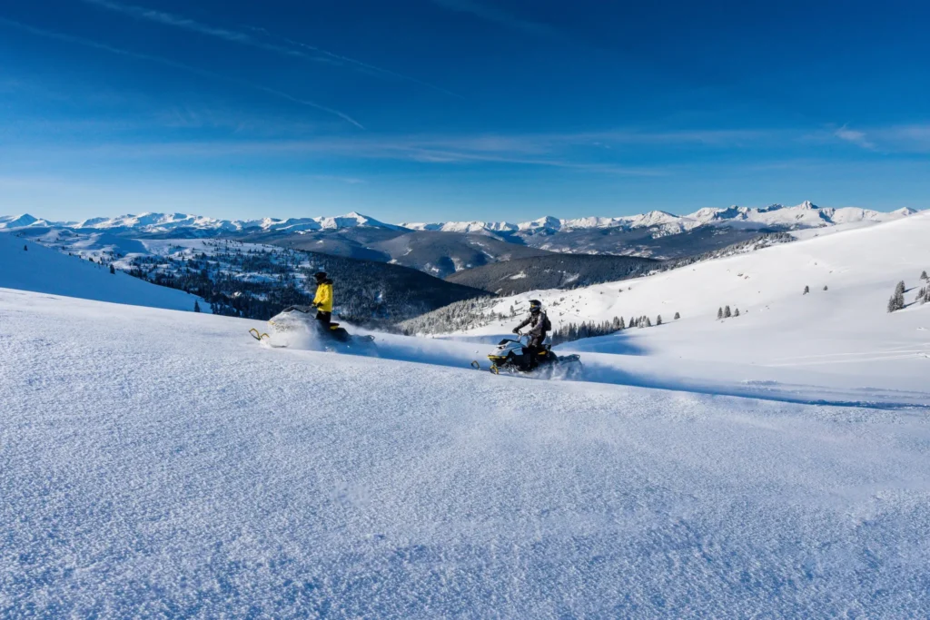 snowmobile riders on snowy mountainside with sweeping views